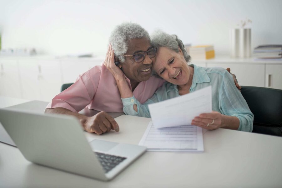 Two joyful senior women embracing while reviewing a document beside a laptop