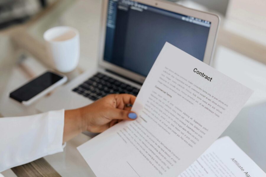 Woman in white coat holding and reading a printed contract document at a desk with a laptop