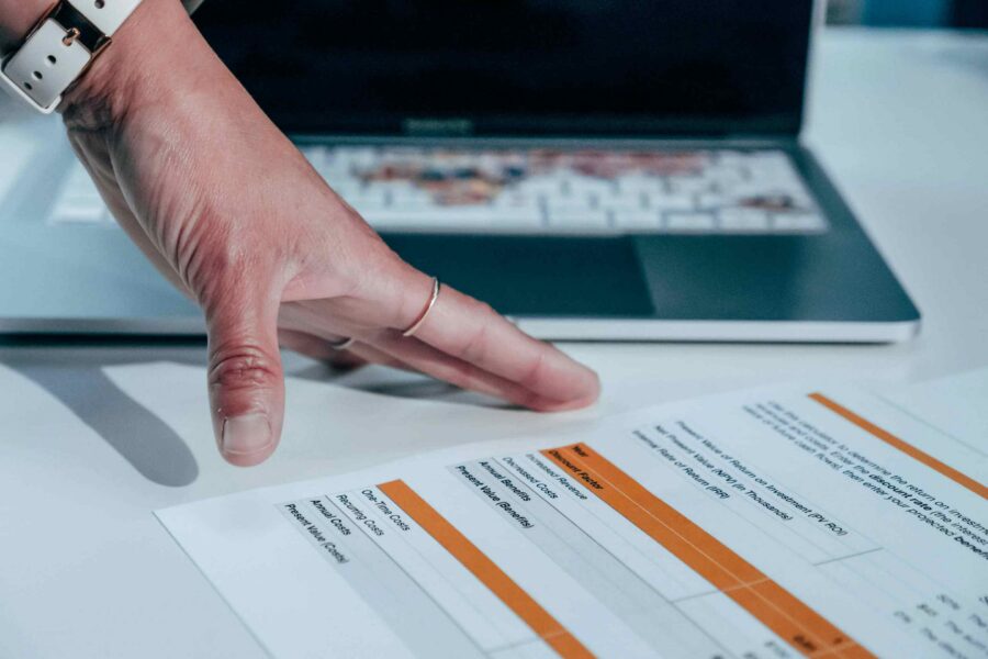 Hand pointing to a printed ROI cost-benefit analysis spreadsheet with orange highlights beside a laptop
