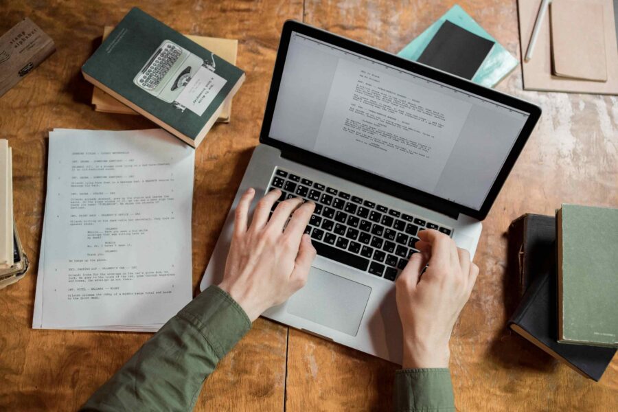 Overhead view of person typing formatted document on laptop surrounded by printed scripts and books