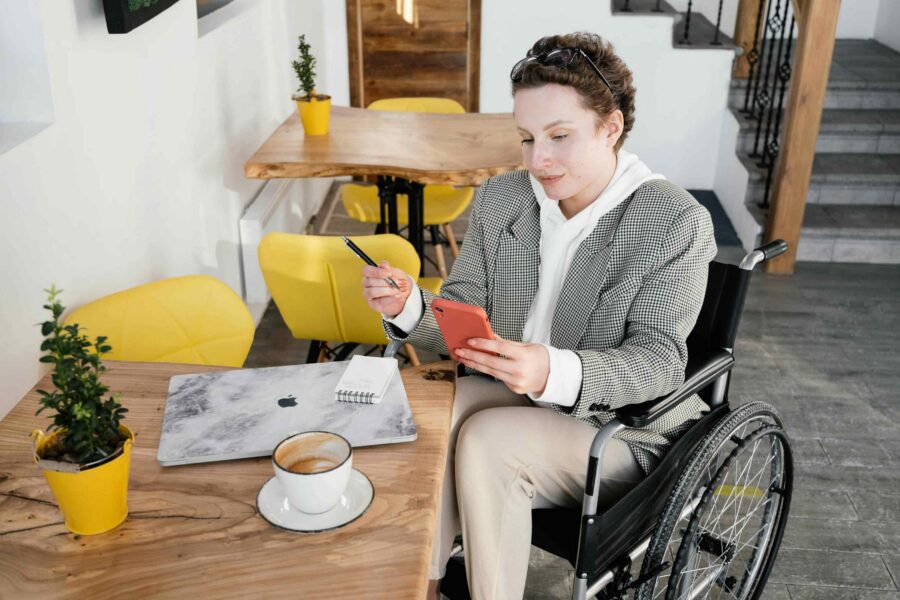 Person in wheelchair using smartphone and laptop at cafe table representing accessible digital document access