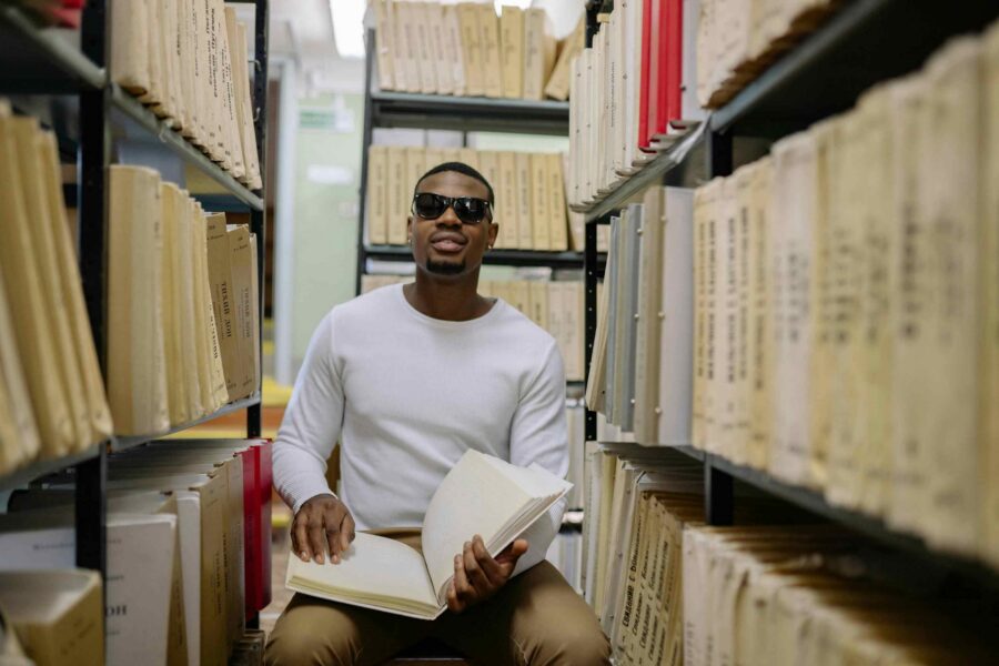 Visually impaired man wearing sunglasses reading open book while seated between library archive shelves
